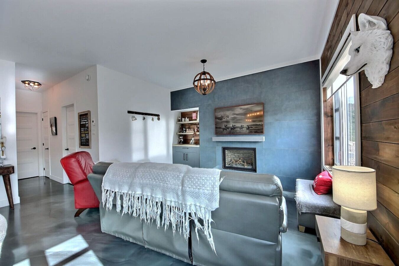 Un salon moderne avec un canapé gris, un fauteuil rouge et un plaid blanc. On y trouve une cheminée surmontée d’un tableau encadré, ainsi qu’une tête d’animal fixée au mur.