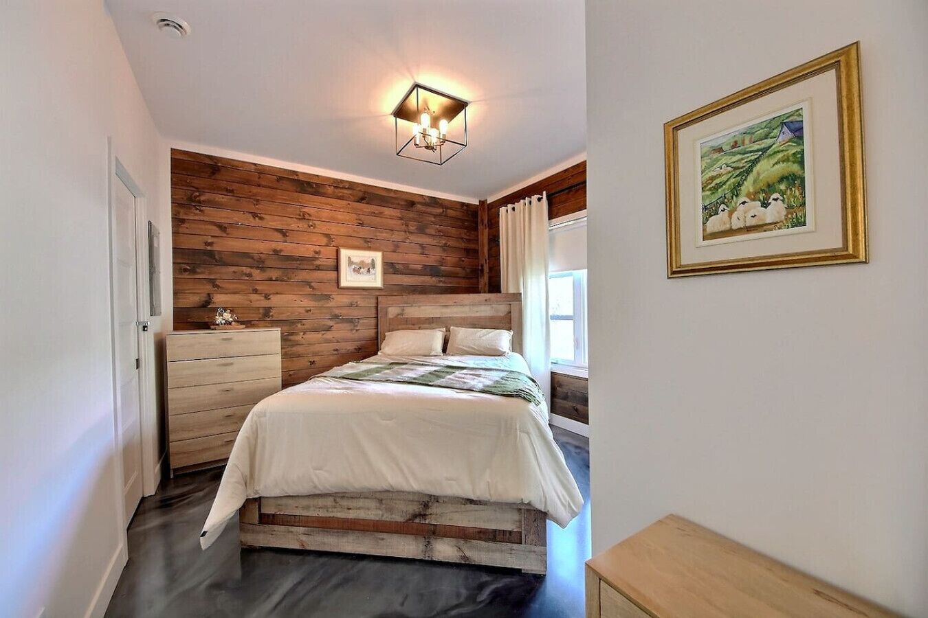 Een slaapkamer met een houten hoofdbord, een bed met witte beddengoed, een nachtkastje, een dressoir en een ingelijst beeld aan de muur.