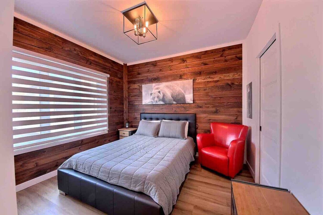 Un dormitorio con cama, una silla roja, paredes de madera, una lámpara en el techo y una ventana con persianas.