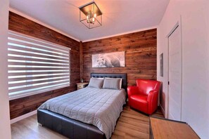 A bedroom with a bed, a red chair, wooden walls, a ceiling light, and a window with blinds. - Condos Monts-Valin- 205, rue du Massif (Valinouët) (Saint-David-de-Falardeau)