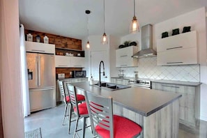 A modern kitchen with a central island, stainless steel appliances, and red bar stools. - Condos Monts-Valin -159, rue du Massif (Valinouët) (Saint-David-de-Falardeau)
