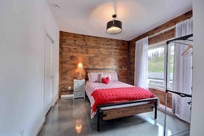 A bedroom with a wooden wall, a bed with a red blanket, a bedside lamp, and a window with curtains. - Condos Monts-Valin -159, rue du Massif (Valinouët) (Saint-David-de-Falardeau)