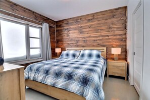 A bedroom with a wooden wall, a bed with a blue and white checkered blanket, two bedside lamps, and a window with curtains. - Condos Monts-Valin - 31, rue du Sommet (Valinouët) (Saint-David-de-Falardeau)