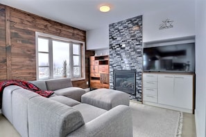 A modern living room with a grey sofa, a fireplace, and a TV. - Condos Monts-Valin - 31, rue du Sommet (Valinouët) (Saint-David-de-Falardeau)