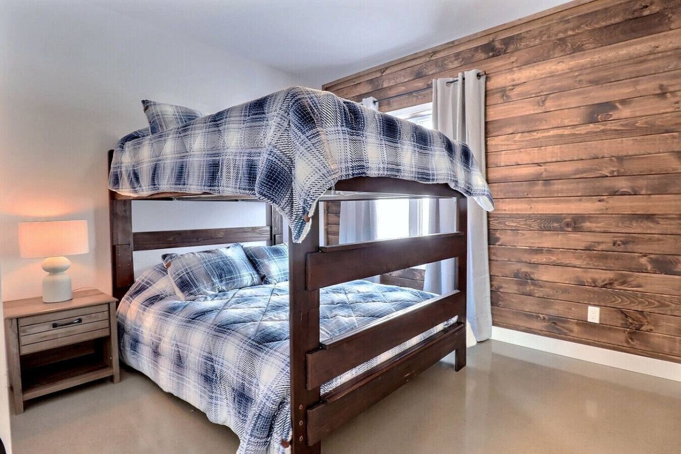 Un lit superposé avec une couverture à carreaux, une table de chevet avec une lampe et un mur en bois.