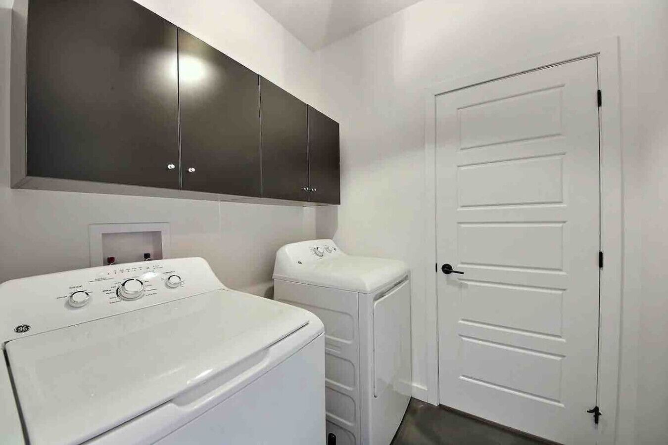 A laundry room with a front-loading washer and dryer, a white door, and dark cabinets above the appliances.