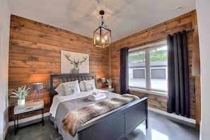 A bedroom with a wooden wall, a bed with a fur throw, a nightstand with a lamp, and a window with curtains. - Condos Monts-Valin -157, rue du Massif (Valinouët) (Saint-David-de-Falardeau)