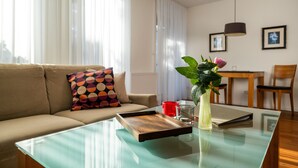 Ein modernes Wohnzimmer mit einem Glastisch, einer Couch mit gemusterten Kissen, einer roten Tasse, einer Vase mit einer Blume und einem Essbereich im Hintergrund.