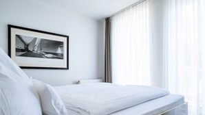 Ein ordentlich bezogenes Bett mit weißen Leinen, ein gerahmtes Foto an der Wand und ein Fenster mit transparenten Vorhängen.