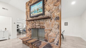 A stone fireplace with a framed painting above it, a stone bench in front, and a kitchen area in the background. - 90+ Day Furnished Retreat! Heated Pool, Spa, Game Room, and 4 King Beds! (Merritt Island)