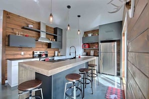 A modern kitchen with a central island, bar stools, pendant lighting, and stainless steel appliances. - Condos Monts-Valin -163, rue du Massif (Valinouët) (Saint-David-de-Falardeau)
