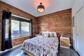 A bedroom with a large bed, wooden walls, a window with a view of a car, and a hanging light fixture. - Condos Monts-Valin -163, rue du Massif (Valinouët) (Saint-David-de-Falardeau)