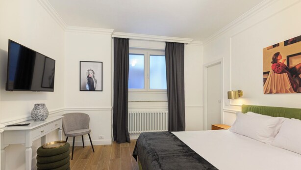 Una camera d'albergo con un letto, una scrivania con televisore, una sedia, un termosifone e una finestra con tende.