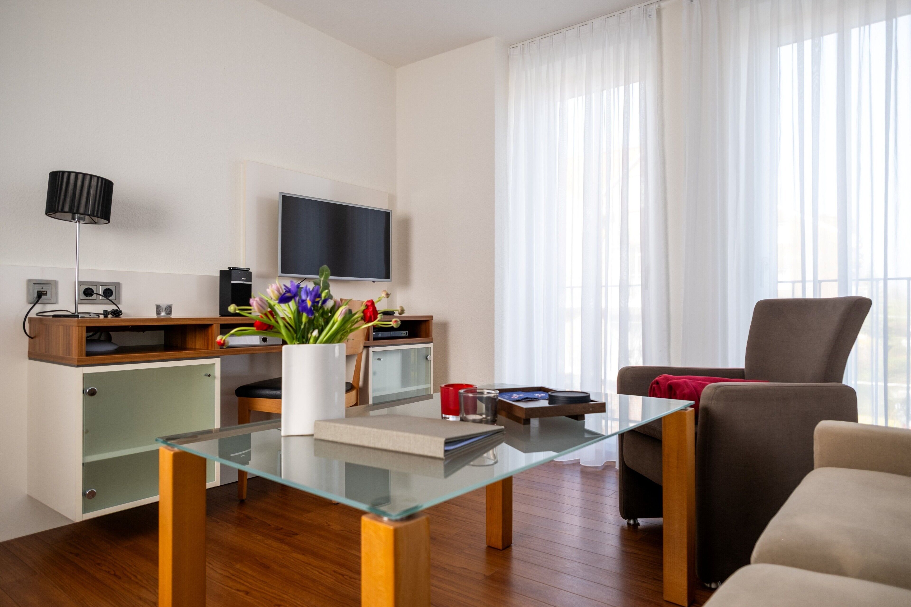 A modern living room with a glass coffee table, a brown sofa, a flat-screen TV mounted on the wall, and a wooden cabinet with a plant.