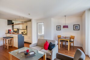 Ein modernes Wohnzimmer mit Glastisch, einer Couch mit Kissen, einem Essbereich mit Tisch und Stühlen, einer Küche mit Herd und Schränken sowie einem Flur mit Spiegel.