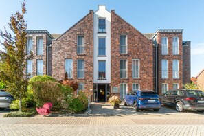 A multi-story brick building with a central entrance, parking area with cars, and a small garden with a red sculpture. - 5-Star Comfort Holiday Home Jümmers - 'Niklas', Prime Location by the Wadden Sea National Park (Wangerland-Horumersiel)