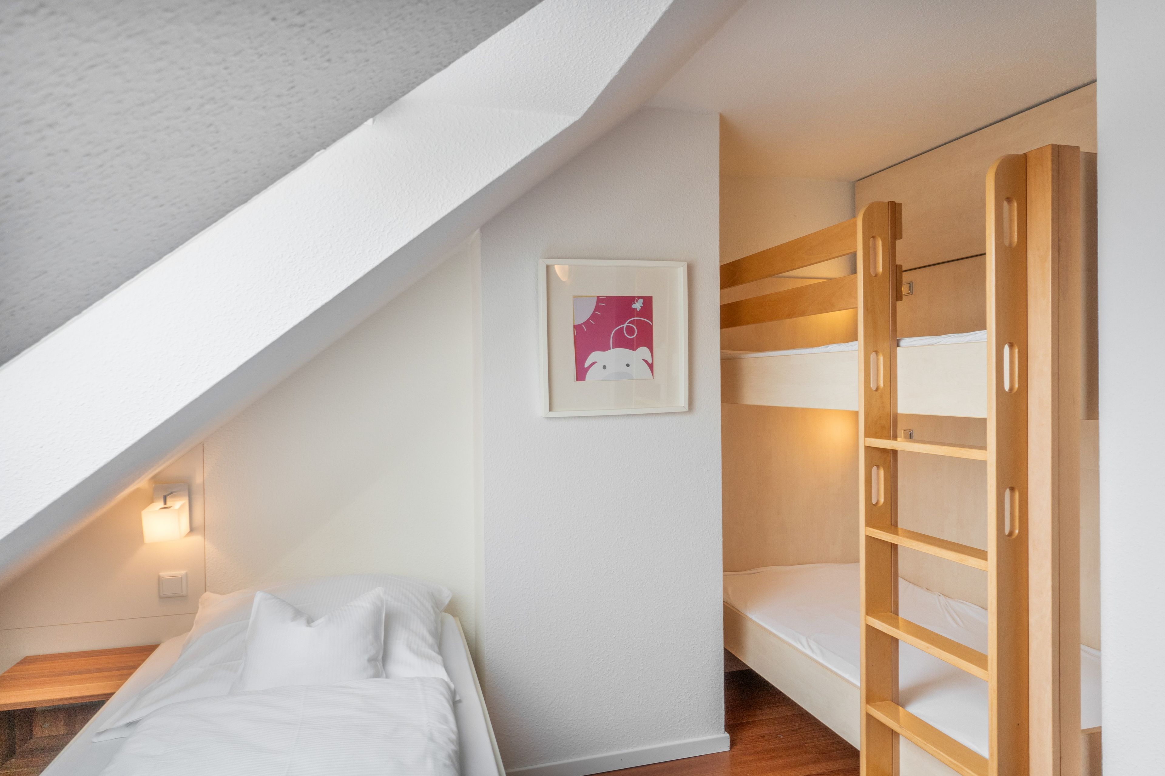 Une chambre avec un lit superposé, un lit simple, un tableau encadré au mur et un plafond en pente.