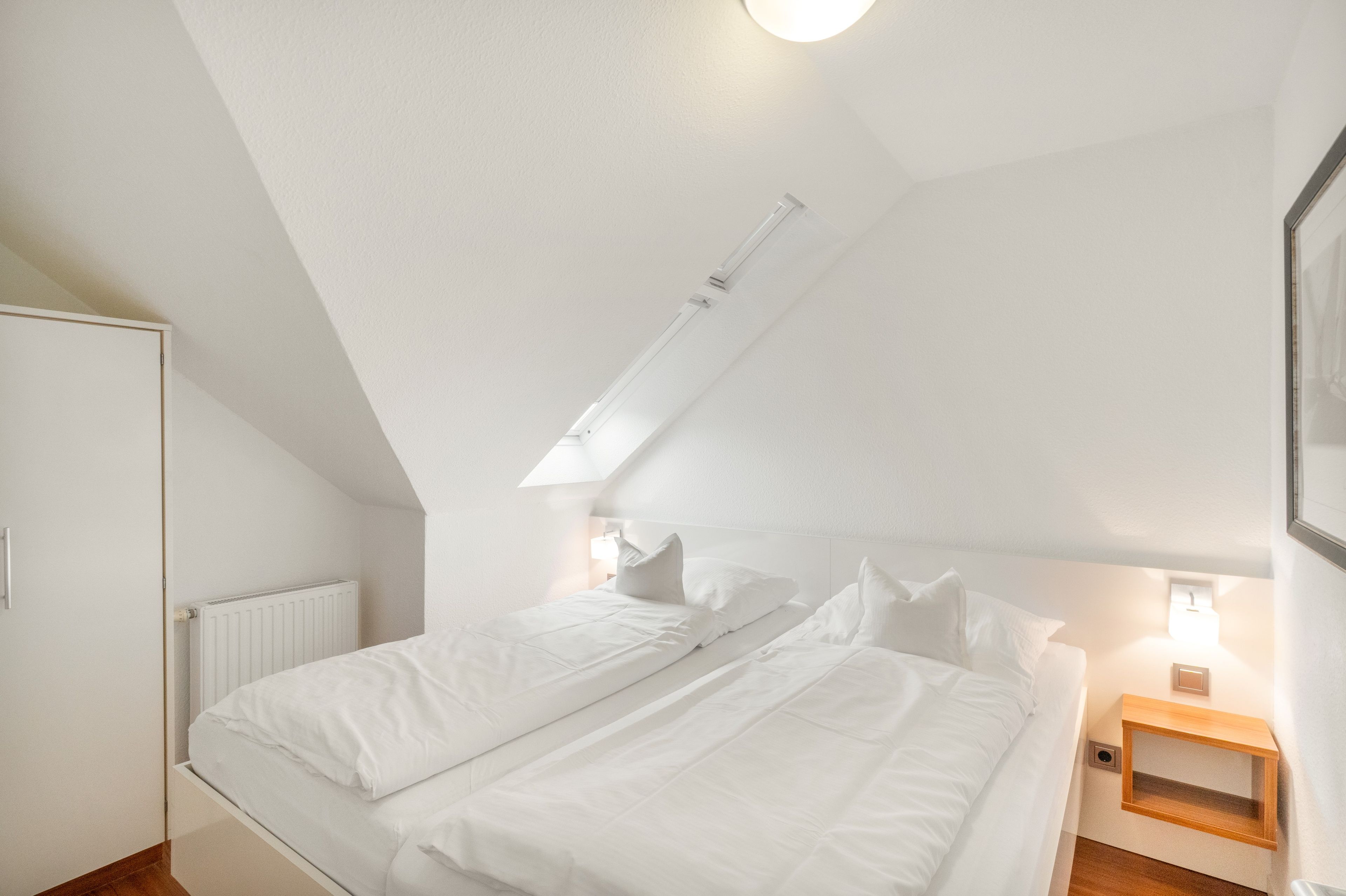 A bedroom with a sloped ceiling, a bed with white bedding, a radiator, and a small wooden nightstand with a lamp.