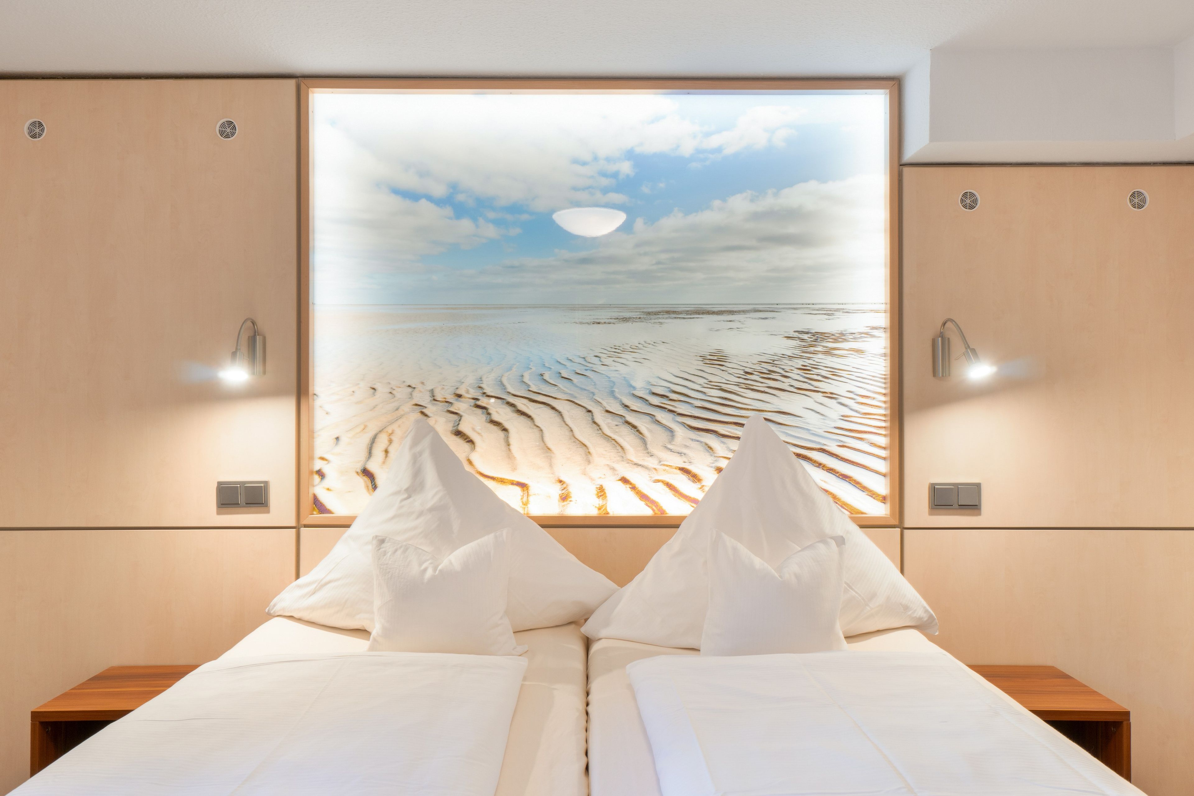 Une chambre à coucher comprenant un lit, deux tables de chevet, une lampe fixée au mur et une grande fenêtre donnant sur la plage.