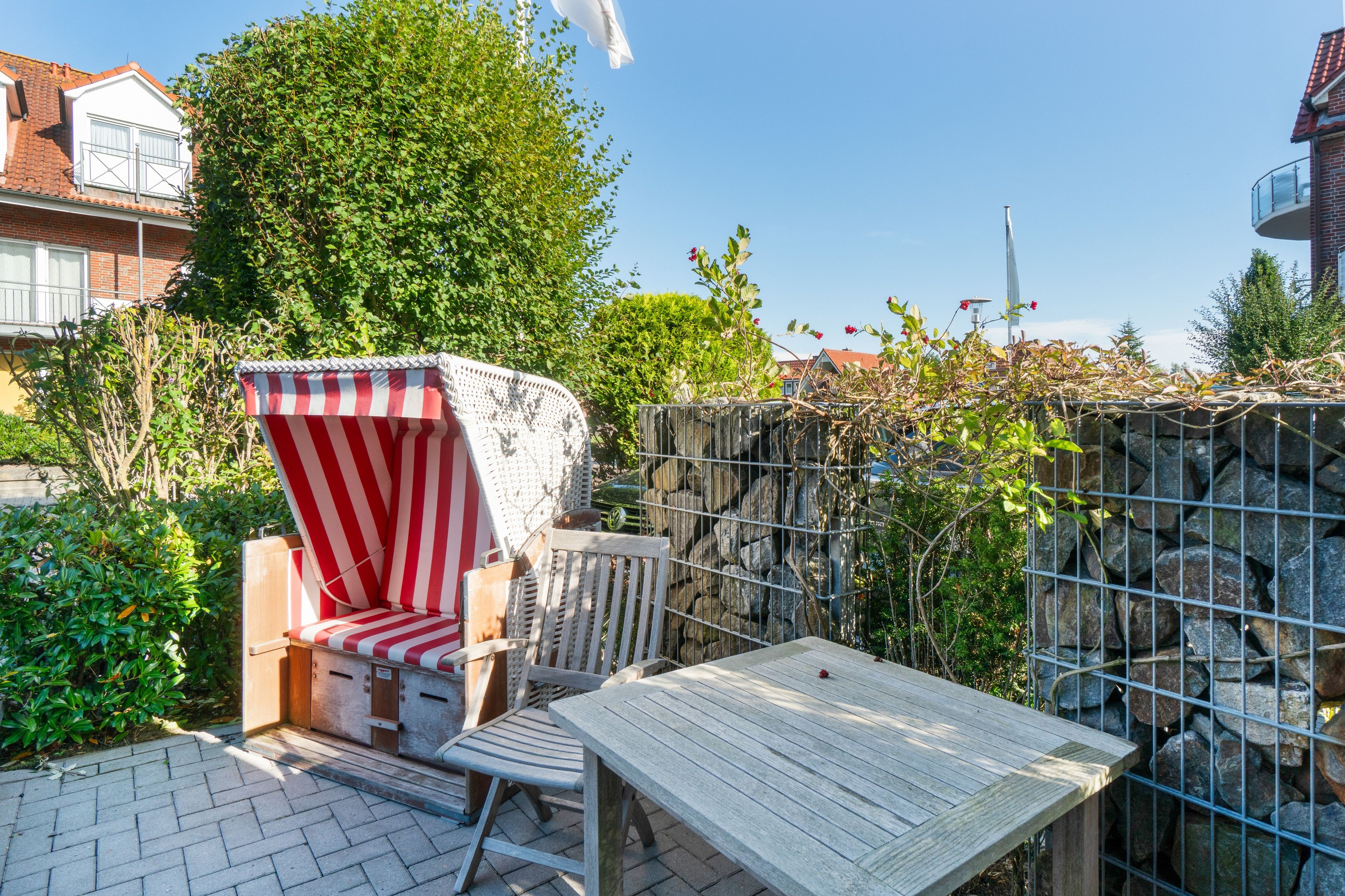 A patio area with a wooden table and a striped beach chair, surrounded by greenery and a stone wall.