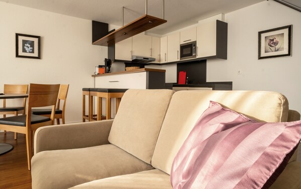 A modern living room with a sofa, a dining table with chairs, a kitchen area with cabinets and appliances, and framed pictures on the walls.