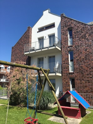 A two-story building with a brick facade, a balcony on the upper floor, and a swing set in the foreground.