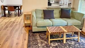 A living room with a green sofa, two pillows, a coffee table, a dining table with chairs, and a kitchen in the background. - Near McKinney Square!  Charming 3 BR house with garage and huge yard (McKinney)