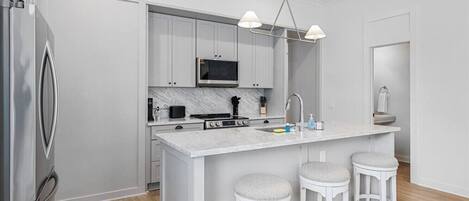 A modern kitchen with white cabinetry, a marble countertop, a built-in oven, and a stainless steel refrigerator. There are three white bar stools and a pendant light fixture above the island.
