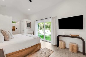 A bedroom with a bed, a TV mounted on the wall, a sliding glass door leading to a balcony, and a small table with stools. - Honey I'm Holmes - New Home Near the Beach! Turf (Holmes Beach)