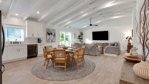 A modern kitchen with white cabinets, a dining area with a round table and chairs, a grey sofa, and a flat-screen TV mounted on the wall. - Honey I'm Holmes - New Home Near the Beach! Turf (Holmes Beach)