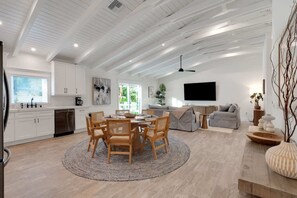 A modern kitchen with white cabinets, a dining area with a round table and chairs, a grey sofa, and a flat-screen TV mounted on the wall. - Honey I'm Holmes - New Home Near the Beach! Turf (Holmes Beach)
