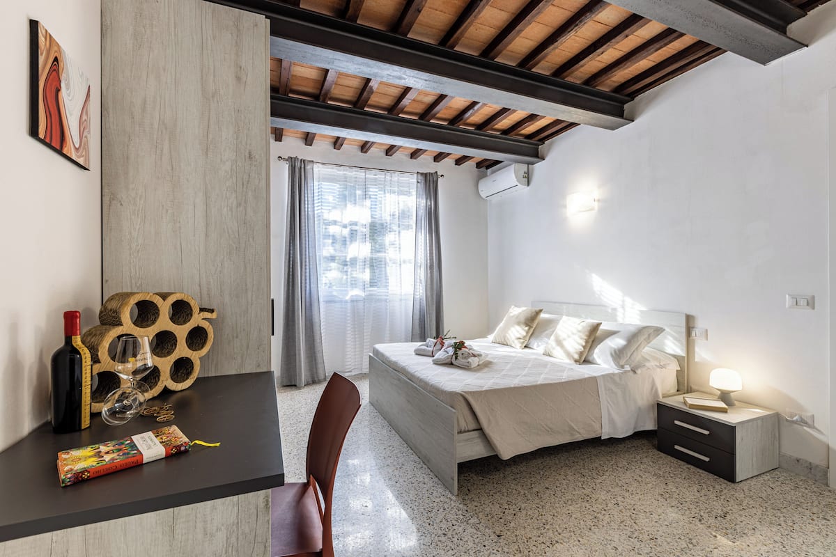 Un dormitorio con cama, mesita de noche y una silla. Se ve una ventana con cortinas, aire acondicionado instalado en la pared y una viga de techo de madera.