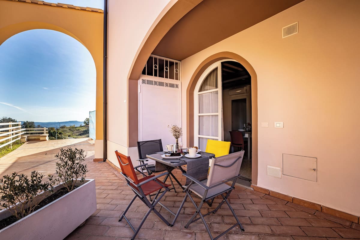 Un patio exterior con una mesa y sillas, una maceta blanca con plantas, y vistas al paisaje.