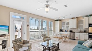 A spacious living area with a glass coffee table, a sofa, and a chair. There is a television on the left and a kitchen area with cabinets and a dishwasher. - Destiny Beach Villas #9B (Destin)
