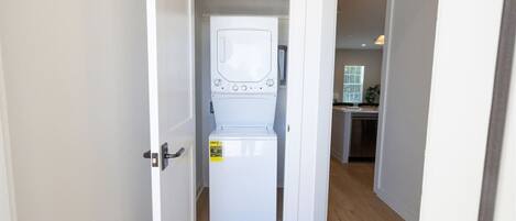 A laundry room with a washing machine and dryer, wooden flooring, and a view into the adjacent kitchen area.