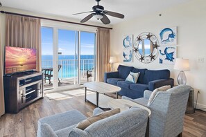 Uma sala de estar com sofá, cadeiras, mesa de centro, TV e varanda com vista para o mar.