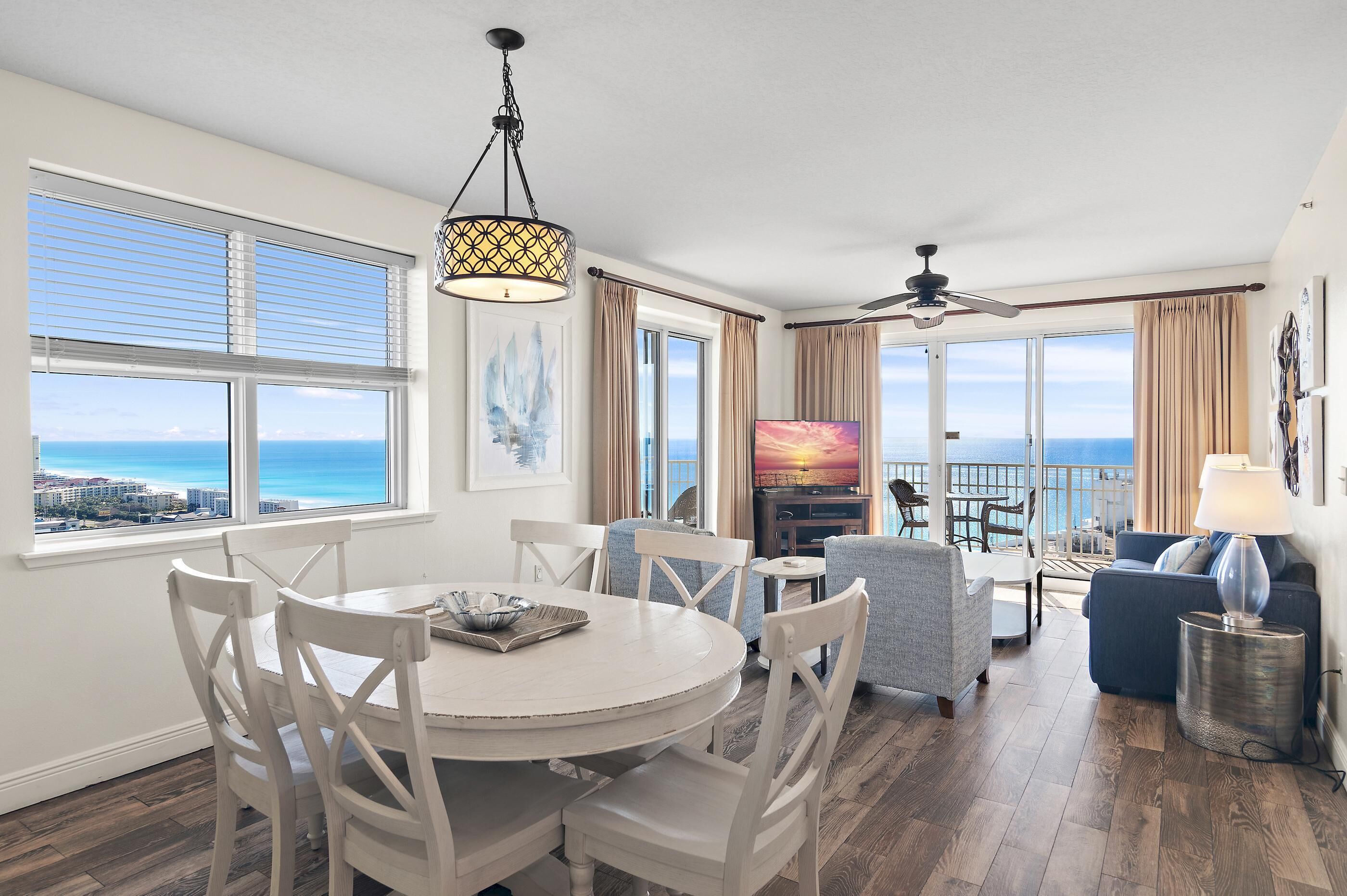 A spacious living area with a dining table, chairs, a sofa, and a TV. Large windows offer a view of the ocean.