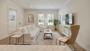 A spacious living room with a white sofa, a coffee table, a TV, and a large window. - Seabreeze Haven 6 - Steps to Beach, Pool, Fast WiFi (Pass Christian)