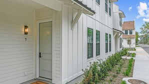 A white two-story residential building with a front door, a small porch, and a well-maintained garden. - Seabreeze Haven 6 - Steps to Beach, Pool, Fast WiFi (Pass Christian)
