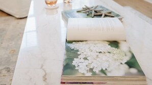 A white marble coffee table with a book, a bottle, and a glass. - Seabreeze Haven 6 - Steps to Beach, Pool, Fast WiFi (Pass Christian)