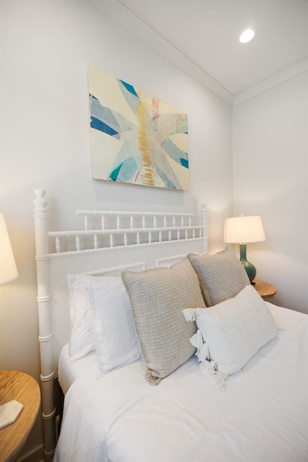 A neatly made bed with white bedding and two pillows, a white headboard, and a wall-mounted abstract painting.