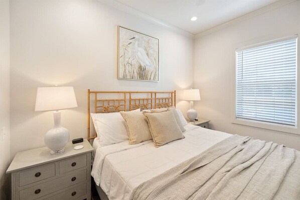 A bedroom with a wooden headboard, white bedding, two bedside tables with lamps, a nightstand, a window with blinds, and a wall-mounted painting of a bird.