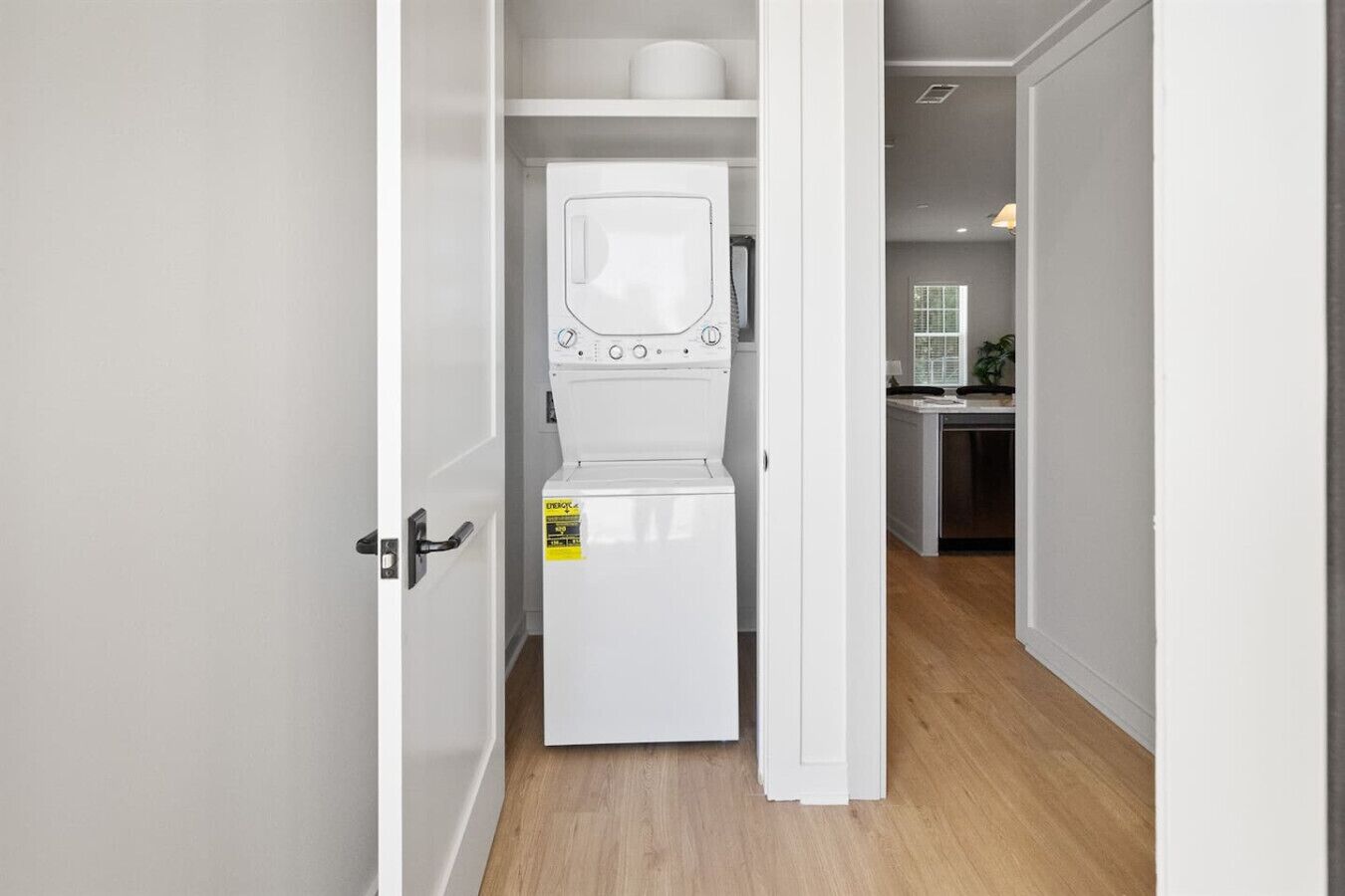 Une petite buanderie équipée d’un lave-linge et d’un sèche-linge à chargement frontal, d’une petite étagère au-dessus du sèche-linge et d’une porte donnant sur un coin cuisine.