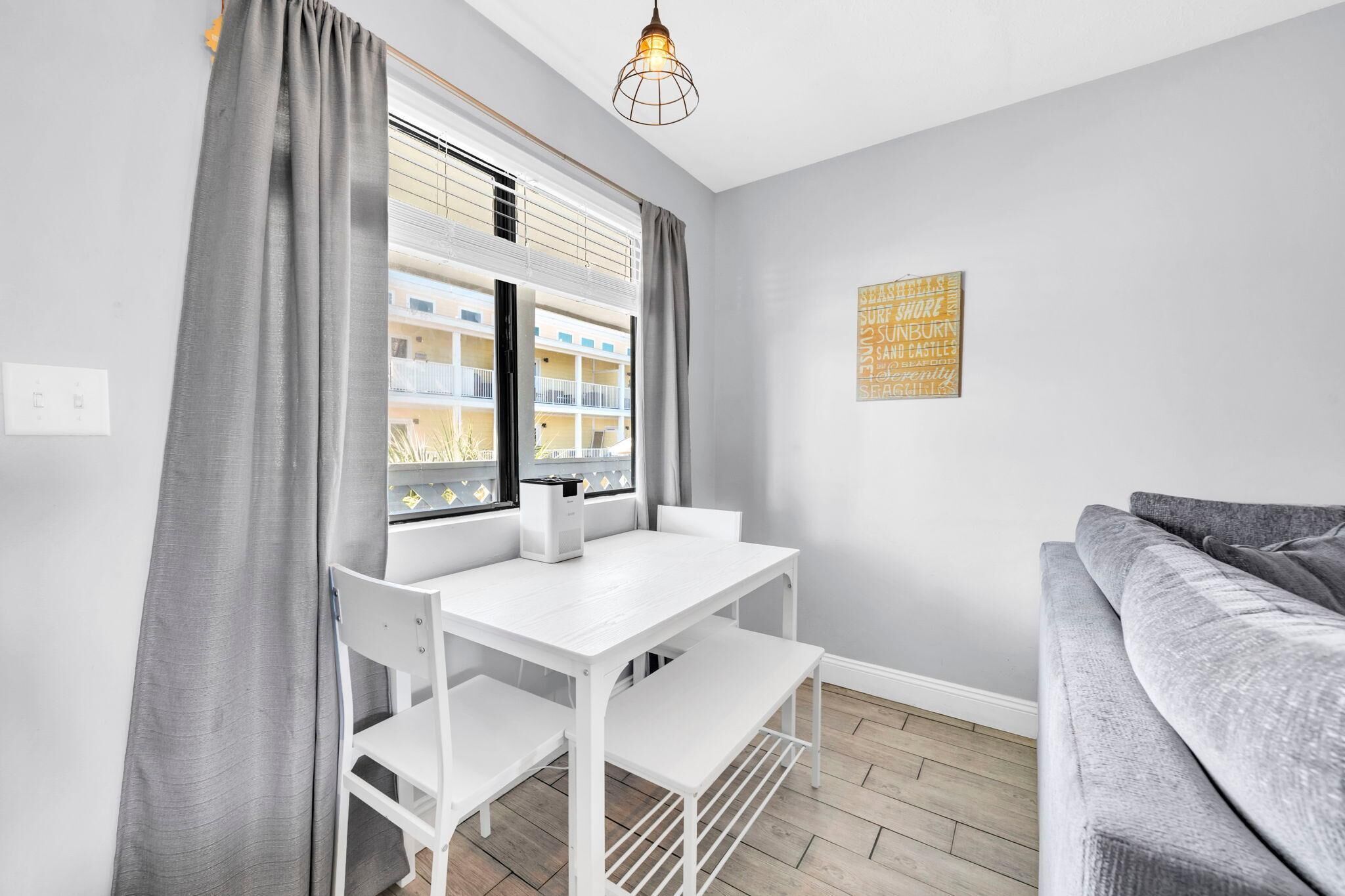 Une pièce moderne avec une table et des chaises blanches, un canapé gris et une fenêtre avec des stores.