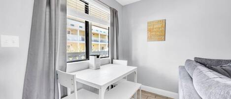 Une pièce moderne avec une table et des chaises blanches, un canapé gris et une fenêtre avec des stores.