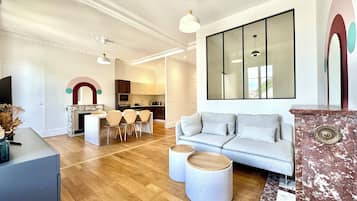 A modern living room with a sofa, a dining area with chairs, a kitchen in the background, and a large mirror on the wall.