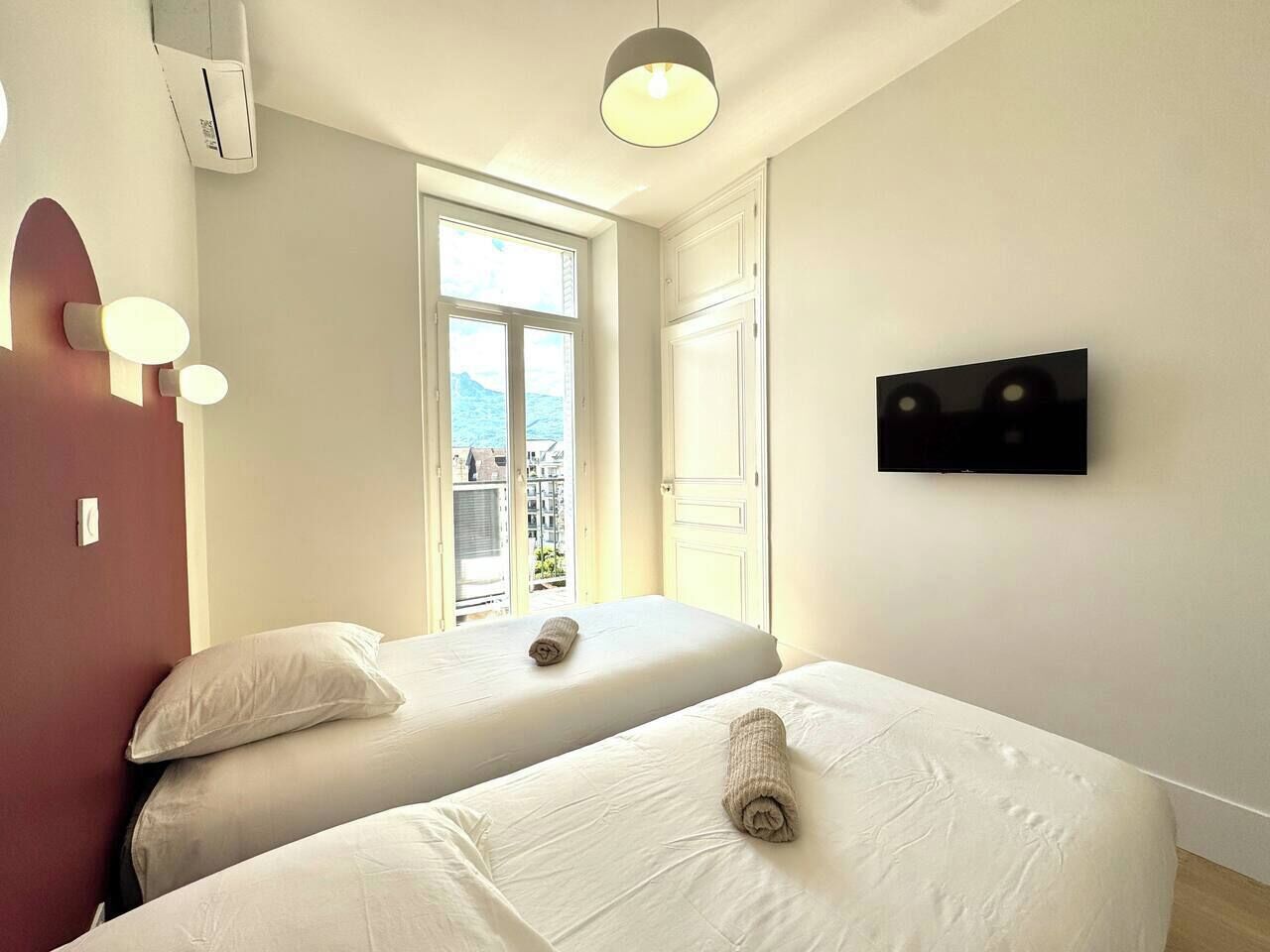 A hotel room with two beds, a TV, and a window with a view of buildings.