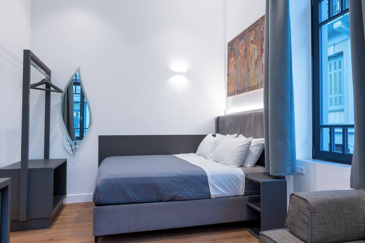 A modern bedroom with a large bed, a side table, a mirror, and a window with blue shutters.