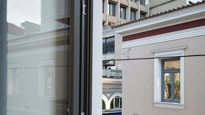 A view from a window overlooking a street with buildings, a balcony, and a shop with outdoor seating. - Lofus Art Suites (Athens)