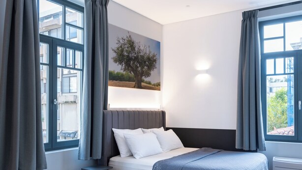A modern bedroom with a large bed, grey curtains, a painting of a tree, and a view of buildings through the window. - Lofus Art Suites (Athens)